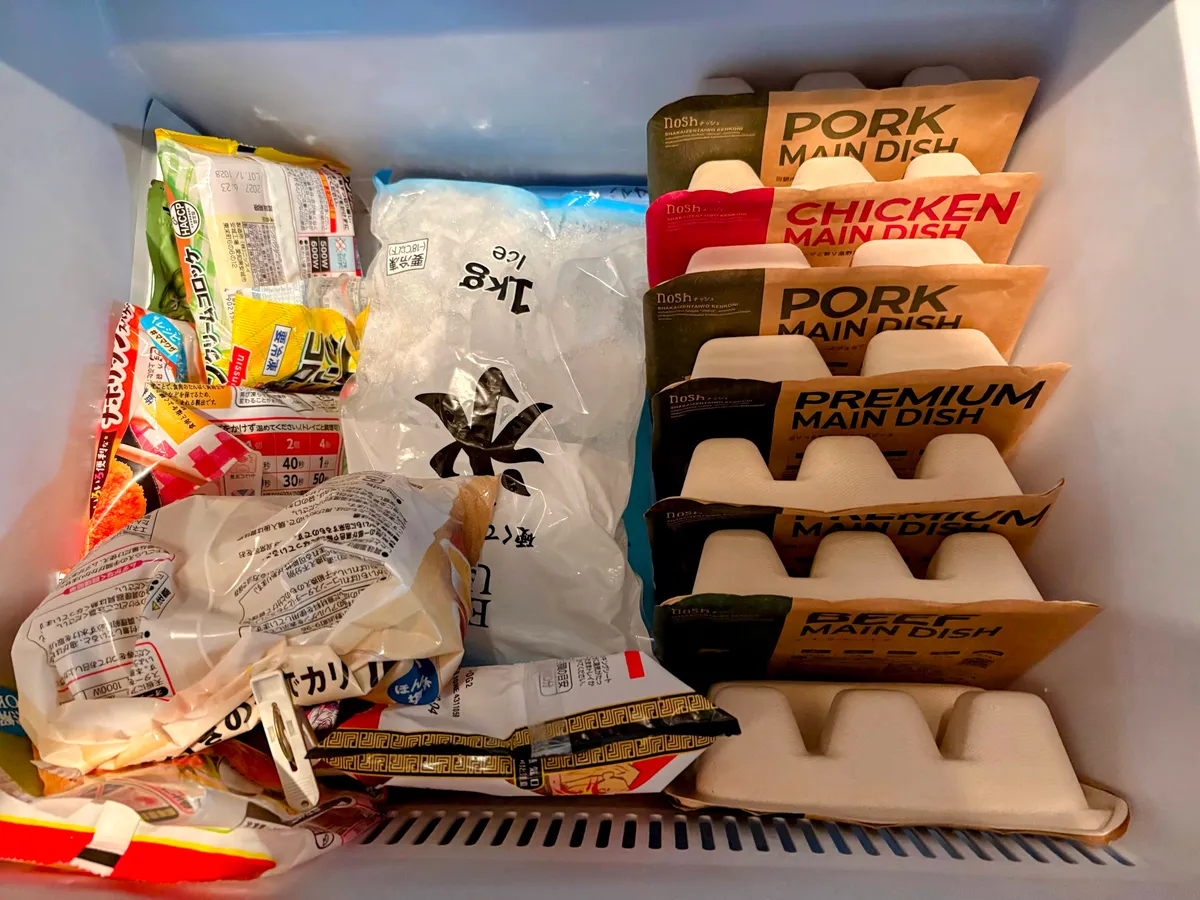 冷凍室の引き出しを上から見た様子。右側の列に宅配食の容器が立てて並び、左側には冷凍麺や冷凍野菜の袋、中央に1kgの氷の袋が入っている。